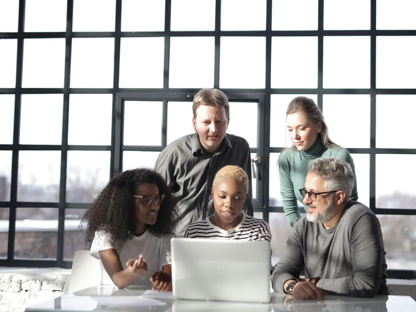 A team crowded around one laptop at a table in front of a tall window